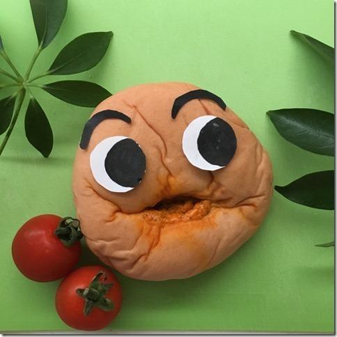 トマトカレーパンの上に目と眉毛が装飾され、中央のくぼみを口に見立てたパンが、2つのミニトマトの横に置かれた写真